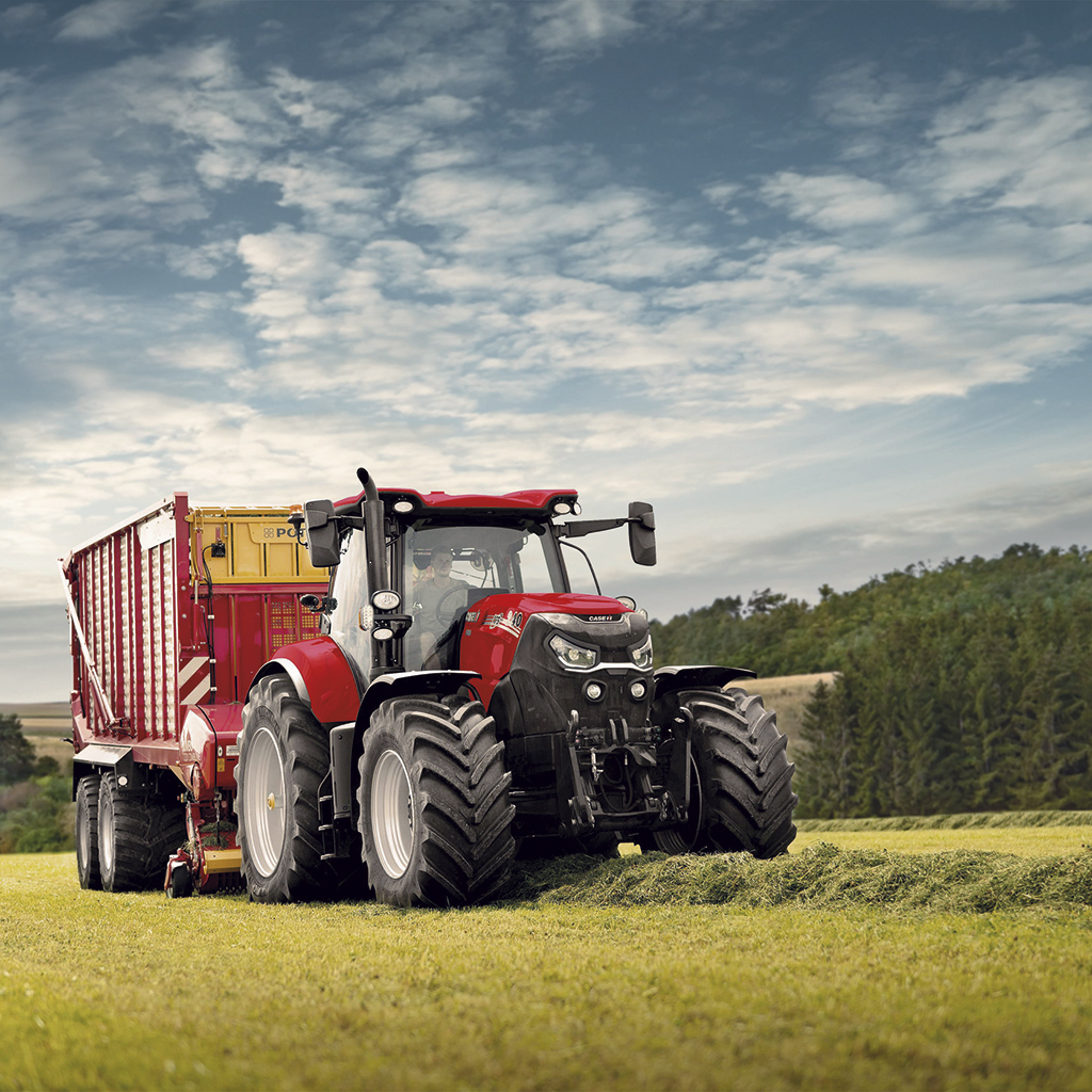 Ein großer, roter moderner Traktor mit Hänger, der frisch gemähtes Gras vor Waldhintergund und wolkigem, blauen Himmel einholt
