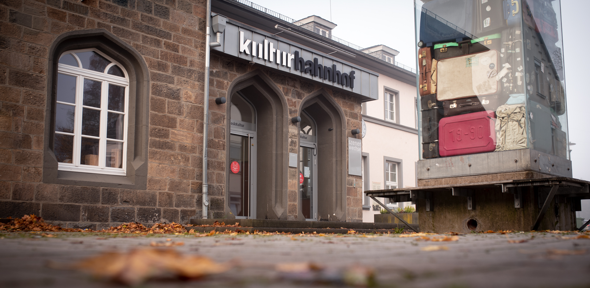 Gebäudeaufnahme des Kreuztaler Kulturbahnhof mit großer Logo-Beschriftung und Koffer-Kunstwerk im Vordergrund in herbstlicher Stimmung.