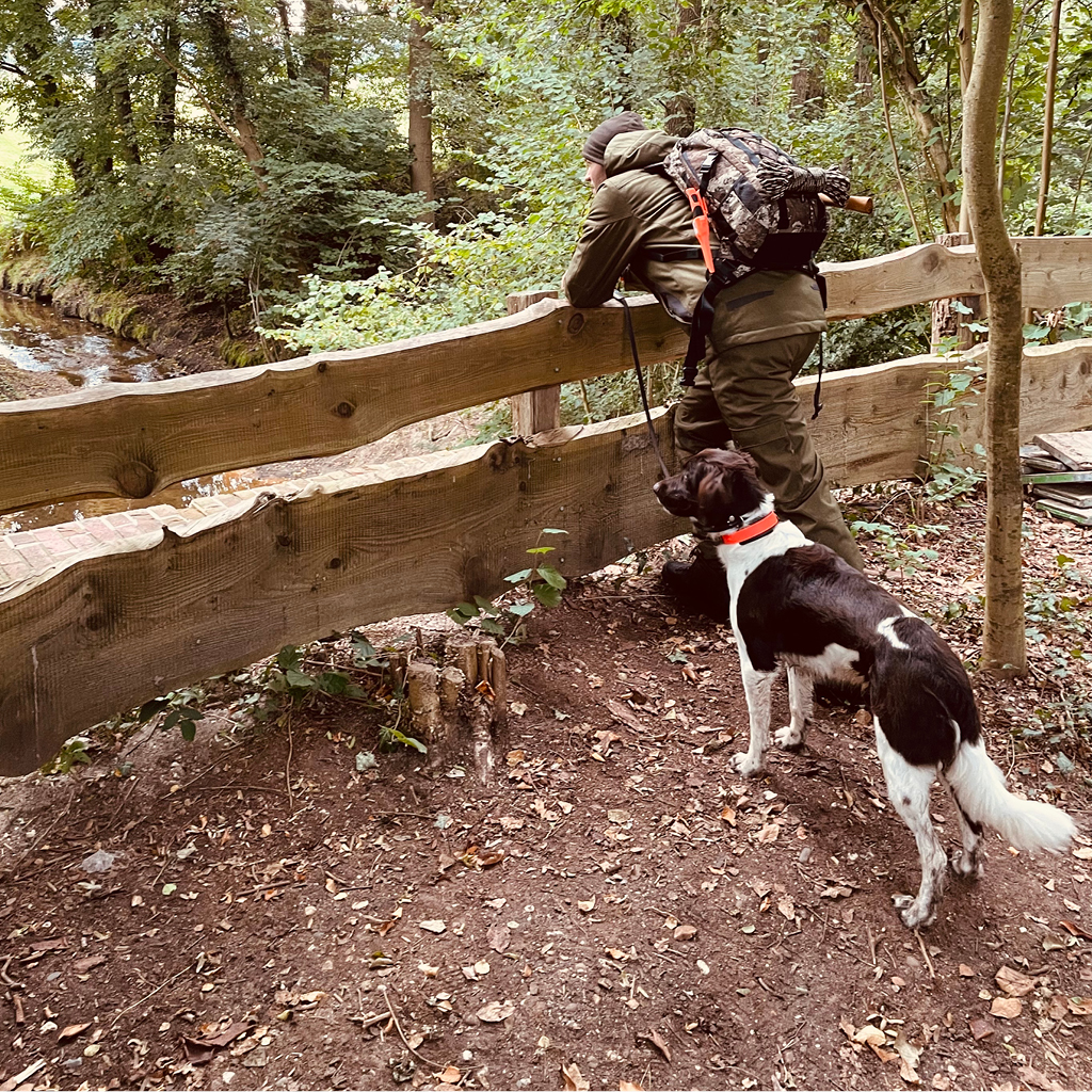 Mann von seitlich hinten mit Outdoorkleidung, Rucksack und Ausrüstung lehnt sich an einen Holzzaun, einen braun-weißen Hund an der Leine. Im Hintergrund Wald und Teil eines Bachlaufes.
