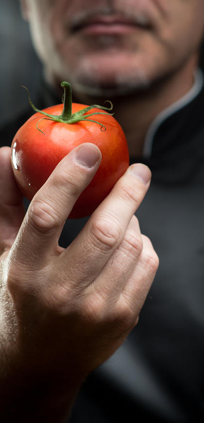 Detailaufnahme einer Tomate mit grünem Stil in der Hand eines Mannes, ein Teil des Gesichtes verschwommen im Hintergrund